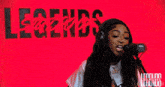 a woman singing into a microphone in front of a red wall with legends written on it