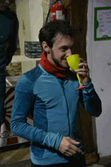a man wearing a blue jacket and a red scarf is drinking from a yellow cup