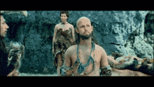a bald man with a beard and a necklace stands in front of a group of people