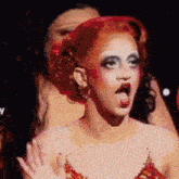 a drag queen with red hair and white makeup is making a funny face with her tongue hanging out .