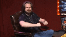 a man with a beard is sitting in a chair in front of a sign that says now .