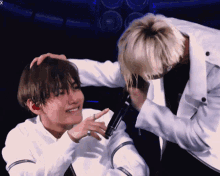 a man in a white jacket is touching another man 's head while holding a microphone