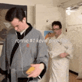 two men in bathrobes are standing next to each other in a room . one of the men is listening to music on his phone .
