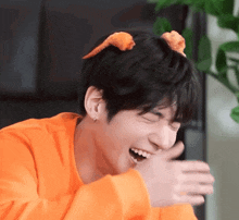 a young man wearing an orange sweater is laughing and covering his mouth .