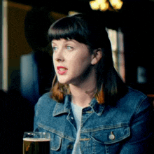 a woman in a denim jacket holds a glass of beer in her hand