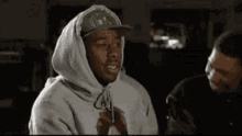 a man in a hoodie and hat is talking to another man in a dark room .