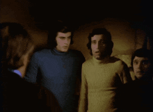 a group of men standing in a dark room with one wearing a blue turtleneck
