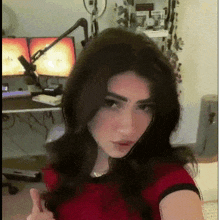 a woman in a red shirt is giving a thumbs up in front of two computer monitors