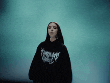 a woman wearing a black hoodie with the word trash on the front stands in front of a blue background