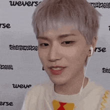 a young man wearing ear buds looks at the camera in front of a wall that says weverse