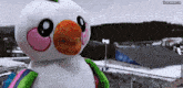 a stuffed snowman with an orange beak and pink cheeks is standing in front of a stadium with a crowd watching a game