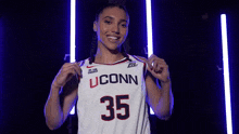 a woman wearing a uconn basketball jersey is smiling and holding her jersey .