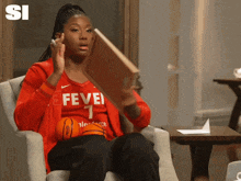 a woman sitting in a chair reading a book while wearing a fever jersey