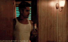 a man in a white tank top is standing in front of a wood paneled wall holding a bottle