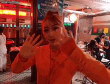 a woman in an orange jacket is giving a peace sign in front of a sign that says honeycam.org