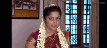 a woman in a red sari is standing in front of a window with a garland of flowers around her neck .