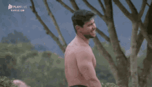 a shirtless man is standing in front of a tree with playplus written on the bottom right corner