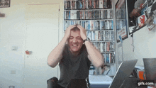 a man is sitting at a desk with his hands on his head in front of a bookshelf full of dvds and a laptop