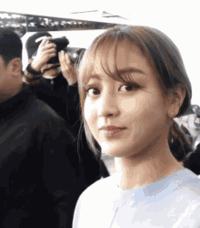 a woman with bangs looks at the camera with a man taking a picture behind her