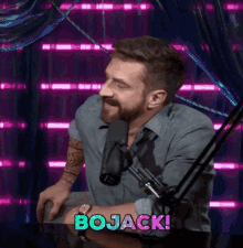 a man with a beard is sitting in front of a microphone and the word bojack is on the screen behind him