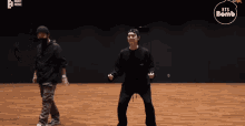 two men are dancing on a wooden floor in a room with a bts bomb logo in the corner .