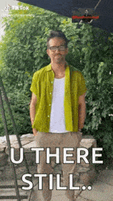 a man in a green shirt is standing in front of a stone wall with his hands in his pockets .
