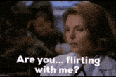 a woman is sitting at a table with a man in the background and says " are you flirting with me "