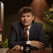 a man in a suit and tie sits at a table next to a mug that says af on it