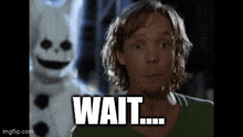 a man in a green shirt is standing in front of a snowman mascot and says `` wait '' .