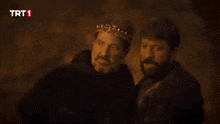 two men are standing next to each other in a dark room and one of them is wearing a crown .