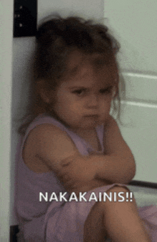 a little girl with her arms crossed is sitting next to a sign that says nakakainis