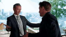 two men in suits and ties are shaking hands in front of a window