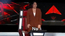 a woman in a suit is standing in front of a red chair on a stage and says `` no '' .