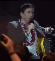 a man wearing a lei is singing into a microphone while sitting on a stage .