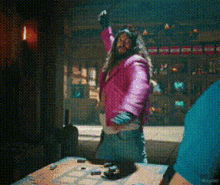 a woman in a pink jacket is standing in front of a table with her arms up .