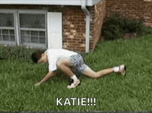 a man is crawling in the grass in front of a brick building and says katie !!!