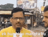 a man wearing glasses and a yellow shirt is talking into a microphone and says ek hug hi to mangta hu .