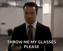 a man in a suit and tie is holding a cell phone and asking to throw me my glasses please .