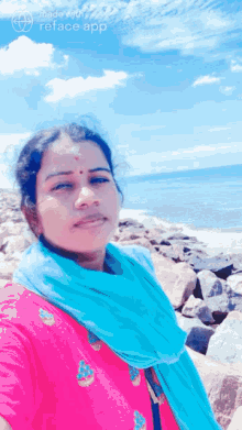 a woman in a pink top and blue scarf stands in front of a beach