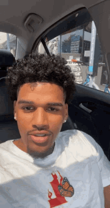 a young man is sitting in a car wearing a white shirt with a butterfly on it .