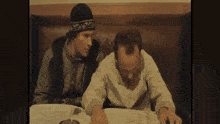 two men are sitting at a table and talking to each other . one of the men is wearing a hat .