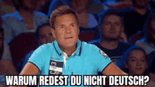 a man in a blue shirt is sitting in front of a crowd and says warum redest du nicht deutsch ?