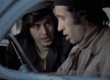 two men are sitting in a car and one is wearing a hat