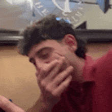a man in a red shirt is covering his mouth with his hand while sitting at a table .