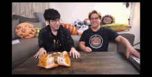 two men are sitting at a table with a bag of food on it