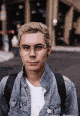 a young man wearing glasses and a denim jacket stands in front of a building