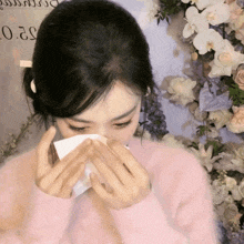 a woman in a pink sweater is blowing her nose in front of flowers