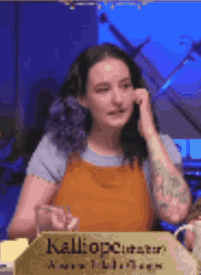 a woman with purple hair and tattoos is sitting at a table with a sign that says kaliopc .