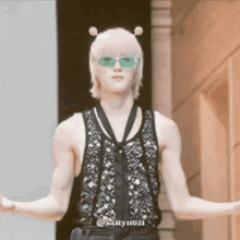 a man wearing a black tank top and green sunglasses is standing with his arms outstretched in front of a building .