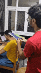 a man in a red shirt is standing next to a man in a yellow shirt sitting at a desk .